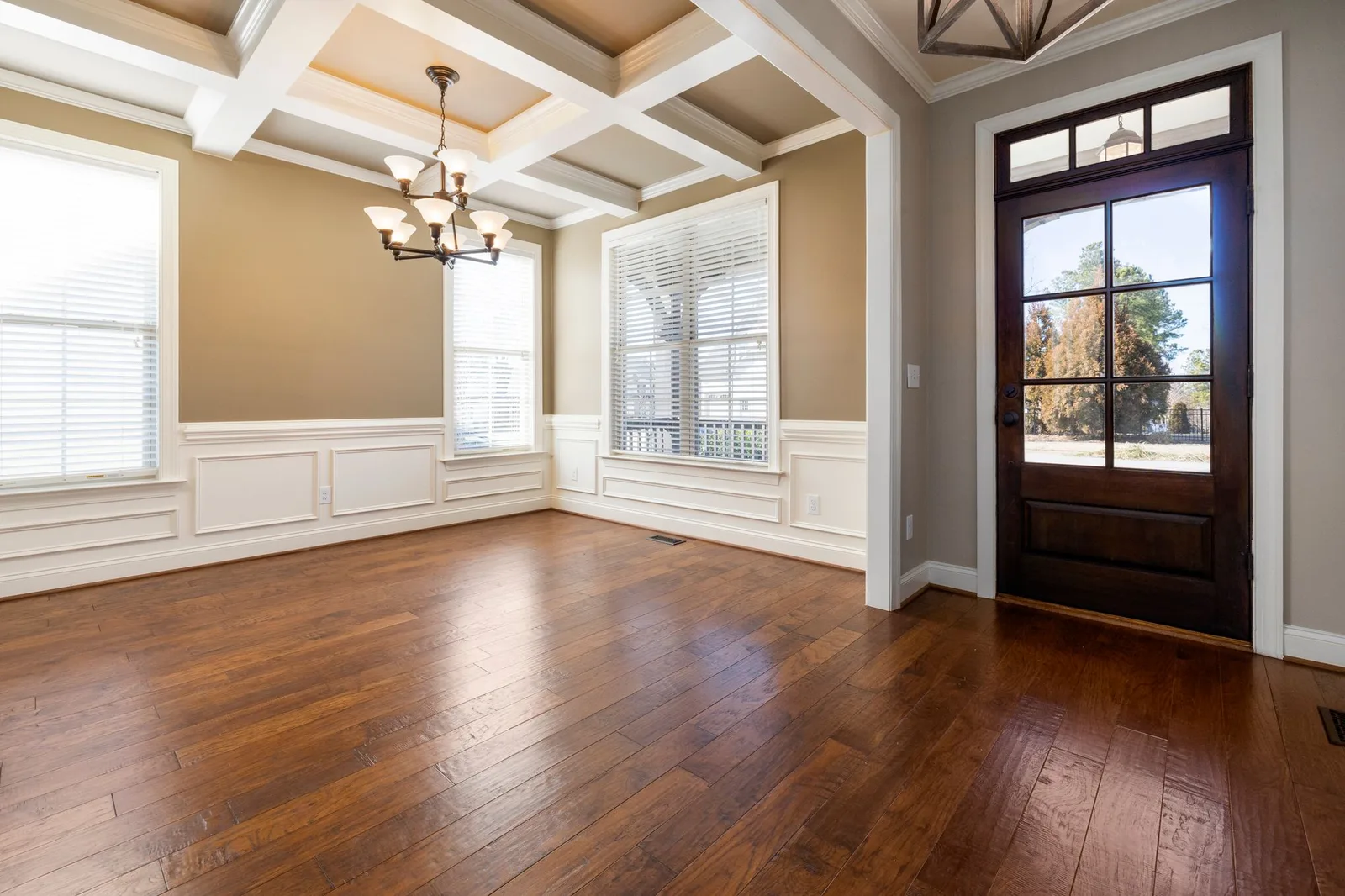 An empty dining room with dark hardwood floors, coffered ceiling and a glass front door.